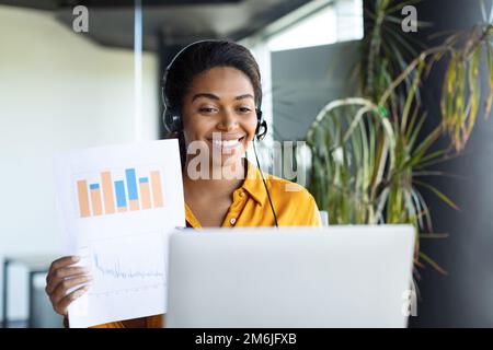 Glückliche schwarze Unternehmerin sitzt am Tisch und zeigt Papier mit Farbtabellen in der Computer-Webcam Stockfoto