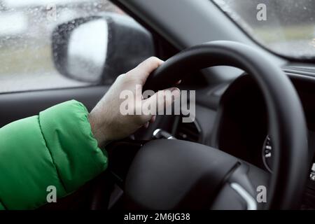 Die Hand eines Mannes hält das Lenkrad eines Autos. Auto fahren an einem Regentag. Weichzeichner. Stockfoto