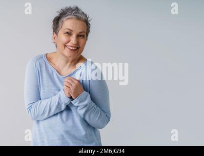 Wunderschöne, reife Frau 50s mit grauen Haaren, die sich posiert, mit Händen nach oben neben dem Herzen und Kopierbereich auf der rechten Seite, isoliert auf weißem Hintergrund Stockfoto