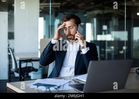 Besorgte männliche Unternehmer, die mit dem Handy telefonieren, den Kopf in Stress berühren und unangenehme Mobiltelefongespräche führen Stockfoto