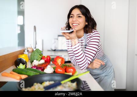 Positive junge araberin, die beim Kochen eine Sprachnachricht aufnimmt Stockfoto
