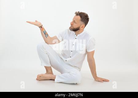 Ruhiger Mann mittleren Alters mit Bart, der mit gebeugten Knien auf dem Boden sitzt und die Hand mit offener Handfläche auf weißem Hintergrund hebt. Stockfoto