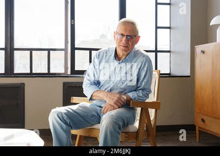 Ein älterer Mann in einer Brille mit Dioptrien sitzt auf dem Stuhl im Wohnzimmer, er schaut in die Kamera Stockfoto