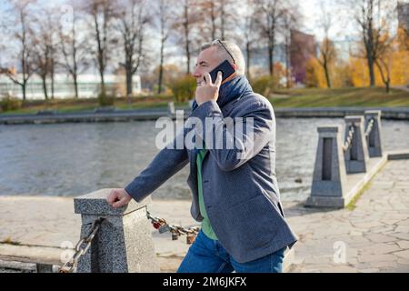 Seitenansicht eines Mannes mittleren Alters, der sich im Herbst auf Betonsäulen lehnt und mit Ketten am Fluss verbunden ist und mit dem Smartphone spricht. Stockfoto