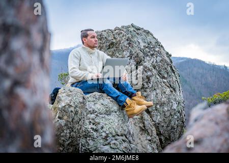 Gut gekleideter Mann, der mit einem Laptop arbeitet, sitzt auf dem felsigen Berg auf dem wunderschönen malerischen clif Hintergrund Stockfoto