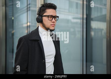 Ein asiatischer Geschäftsmann mit Kopfhörern läuft am Bürogebäude vorbei. Stockfoto