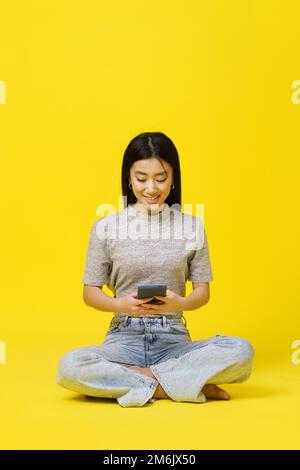 Asiatisches junges Mädchen, das auf dem Boden sitzt und mit einem Smartphone Textnachrichten schickt oder online einkauft, isoliert auf gelbem Hintergrund. Produktplatz Stockfoto