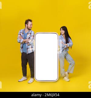 Asiatisches Mädchen und weißer Kerl, der stand, lehnte sich an ein riesiges Smartphone mit weißem, leerem Bildschirm, Werbung für mobile Apps und Lächeln-Look Stockfoto