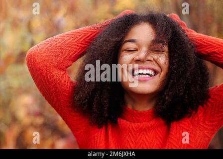 Junge, glückliche, lockige afroamerikanische Frau mit geschlossenen Augen und lachend im Herbstwald Stockfoto