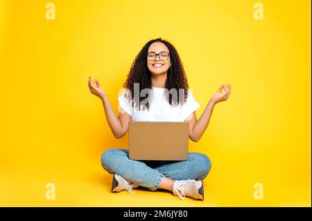 Foto einer positiven, reizenden Latino- oder brasilianischen Frau in Freizeitkleidung, die mit einem offenen Laptop in Lotusposition sitzt und die Augen auf einem isolierten gelben Hintergrund geschlossen hat, sich entspannt und meditiert Stockfoto