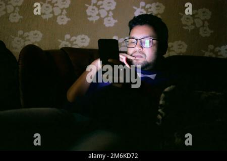 Junger Mann, der nachts mit einem Smartphone auf dem Bett sitzt Stockfoto
