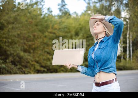 Porträt einer jungen, selbstbewussten blonden Frau, die unbeschriftete Pappe hält, die Hand hebt, um die Augen vor der Sonne zu verdecken, und eng aussieht. Stockfoto