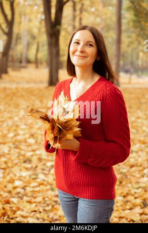 Porträt einer jungen, makellosen, lächelnden Frau, die zwischen gelben gefallenen Blättern im Wald steht, einen Blattrand hält und posiert. Stockfoto