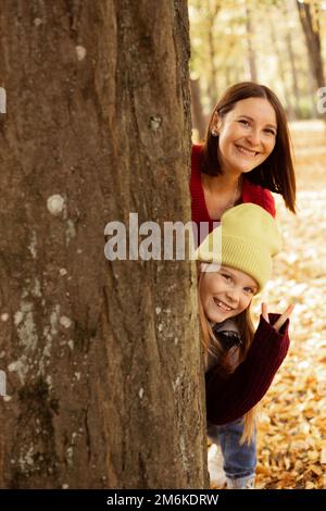 Porträt einer Familie von jungen Mutter, Teenager, die hinter einem Baum steht, mit gelben, gefallenen Blättern, die aus dem Kofferraum schauen. Stockfoto