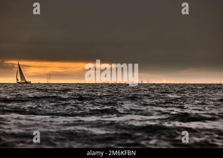 Segelboot im Meer bei stürmischem Wetter, stürmische Wolken Himmel orange, Segelregatta, Reflexion des Segeles im Wasser, bigl Wellen von wa Stockfoto