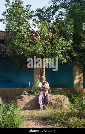 NOVEMBER 15 2022, KACHANDA, CHHATTISGARH, INDIEN: Großvater hält den Enkel im indischen Dorf und sitzt im Morgenlicht unter dem Baum. Großvater Stockfoto