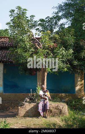 NOVEMBER 15 2022, KACHANDA, CHHATTISGARH, INDIEN: Großvater hält den Enkel im indischen Dorf und sitzt im Morgenlicht unter dem Baum. Großvater Stockfoto