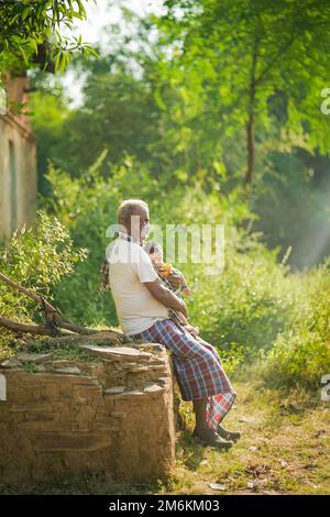 NOVEMBER 15 2022, KACHANDA, CHHATTISGARH, INDIEN: Großvater hält den Enkel im indischen Dorf und sitzt im Morgenlicht unter dem Baum. Großvater Stockfoto