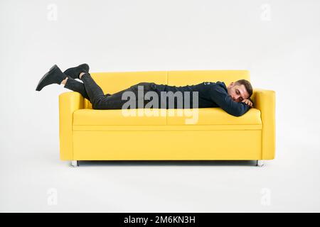 Ein müder junger Mann, der auf einem gelben Sofa auf weißem Hintergrund liegt Stockfoto
