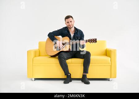 Attraktiver junger Mann, der Akustikgitarre spielt, sitzt auf einem gelben Sofa auf weißem Hintergrund Stockfoto