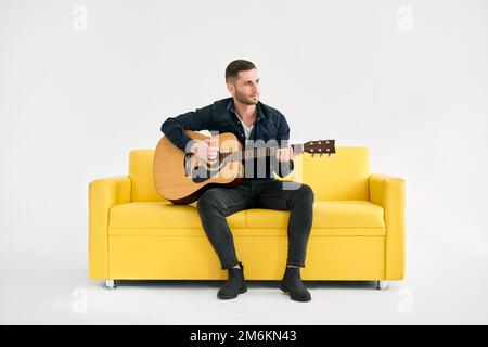 Attraktiver junger Mann, der Akustikgitarre spielt, sitzt auf einem gelben Sofa auf weißem Hintergrund Stockfoto
