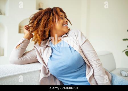 afroamerikanisches Millennialmädchen, zufrieden und glücklich, sitzen auf dem bequemen Sofa in einem gemütlichen, modernen, hellen Wohnzimmer Stockfoto