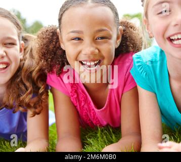 Sie hat immer Spaß mit ihren besten Freunden. Drei multiethnische junge Mädchen, die auf dem Rasen in einem Park lagen und vor der Kamera lächelten. Stockfoto