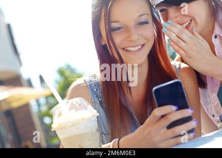 Einen unartigen Text teilen. Eine Teenagerin, die SMS auf einem Handy schreibt, während ihre Freundin ihr etwas ins Ohr flüstert. Stockfoto