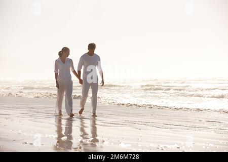 Spazieren am Strand mit jemand Besonderem. Aufnahme eines attraktiven jungen Paares in Weiß, das am Strand entlang spaziert. Stockfoto