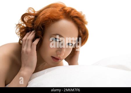 Lächelnde rothaarige junge Frau, die im Bett liegt und sich unter dem Kissen versteckt Stockfoto