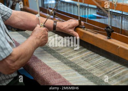 Ein Meisterhandwerker, der an einem traditionellen Holzwebel arbeitet, und ein handgewebtes Wollgewebe mit Fischgrätmuster Stockfoto