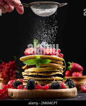 Ein Stapel Pfannkuchen mit frischem Obst, bestreut mit Puderzucker auf schwarzem Hintergrund Stockfoto