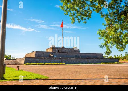 Atemberaubender Blick auf einen Platz und die Flagge von Vietnam (rote Flagge mit goldenem Stern), die über einem Turm der Zitadelle auf blauem Himmel in Hue, Vie flattert Stockfoto