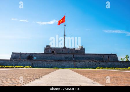 Atemberaubender Blick auf einen Platz und die Flagge von Vietnam (rote Flagge mit goldenem Stern), die über einem Turm der Zitadelle auf blauem Himmel in Hue, Vie flattert Stockfoto