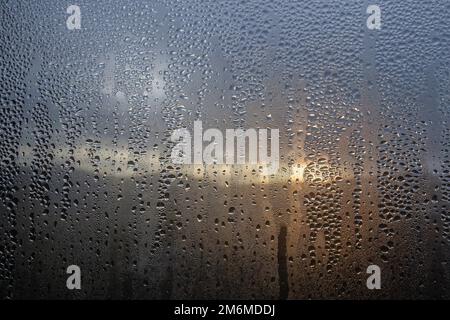 Kondensation am Fenster mit stürmischem Sonnenaufgang draußen, Wassertropfen auf dem Glas. Stockfoto