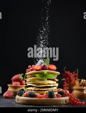 Ein Stapel Pfannkuchen mit frischem Obst, bestreut mit Puderzucker auf schwarzem Hintergrund Stockfoto