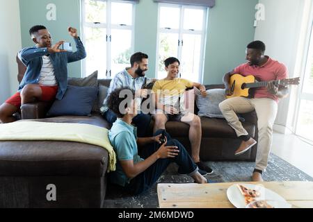 Multirassische Freunde, die Bier genießen und Smartphone benutzen, während sie jungen Mann beim Gitarrenspiel betrachten Stockfoto