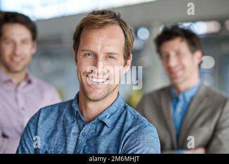 Hervorragende Arbeitsbeziehungen unter Kollegen. Ein gutaussehender Mann lächelte in die Kamera, mit zwei männlichen Kollegen hinter ihm. Stockfoto