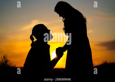 Die Silhouette eines Mannes, der vor einer Frau kniet, vor dem Hintergrund eines wunderschönen, ruhigen Sonnenuntergangs - Verlobungskonzept Stockfoto