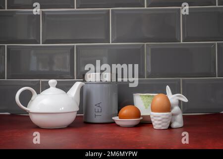 Frühstück mit gekochten Eiern in Eierbecher und Tasse Tee mit Caddy und Teetasse und Untertasse auf einem roten Linoleum Arbeitsplatte Stockfoto