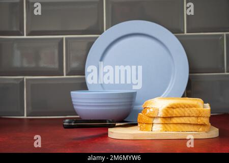 Blaue Keramik essteller Schüssel Edelstahlmesser mit schwarzem Griff auf Holzbrett mit frischem Brot auf rotem Linoleum Arbeitsplatte mit grauen Fliesen Stockfoto