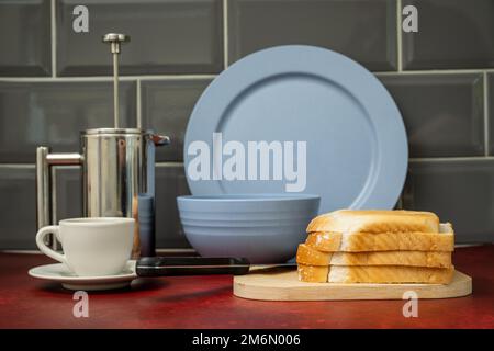 Espresso-Kaffeetasse und -Untertasse Edelstahl cafetiere schwarzes Edelstahlmesser auf Holzbrett mit frischem Brot blaues Abendset auf Rot Stockfoto