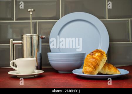 Espresso-Kaffeetasse und Untertasse aus Edelstahl cafetiere mit frischem Coissant Blue Dinner-Set auf rotem Linoleum Arbeitsplatte mit grauem Fliesen auf der Rückseite Stockfoto