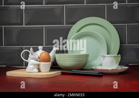 Zum Frühstück gekochtes Ei in einem porzellan-Eierbecher mit porzellan-Teekanne und Untertasse und grünem Geschirr auf roter Arbeitsplatte und grauen Fliesen Stockfoto