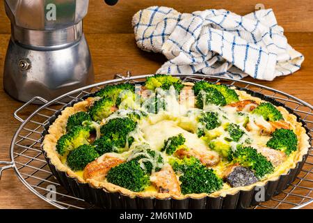 Frisch gebackener Brokkoli und hausgemachter Lachs Quiche in schwarzer Tortenpfanne auf Metallkühlregal mit Moka-Kanne Kaffeemaschine auf Holztisch. Stockfoto