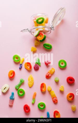 Overhead-Ansicht der verstreuten fallen bunten Zucker Süßigkeiten mit offenem Glas auf rosa Hintergrund Stockfoto