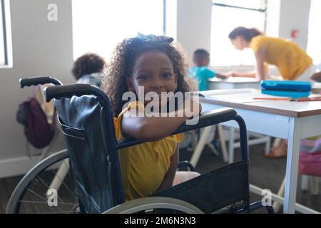 Porträt eines lächelnden afroamerikanischen Grundschulmädchen, das im Klassenzimmer auf dem Rollstuhl am Schreibtisch sitzt Stockfoto