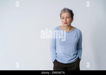 Gestresste, wütende, reife, grauhaarige Frau 50s, die sich als frustriert ausgibt und mit den Händen in den Taschen auf die Kamera schaut, Kopierraum auf der linken isola Stockfoto