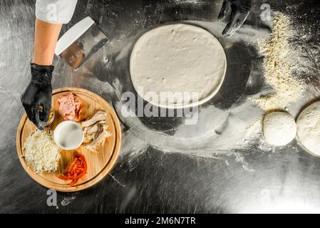 Pizza auf einem Stahltisch kochen; Teig, Mehl, Pizza-Belag, Sauce und Hände des Küchenchefs in schwarzen Handschuhen auf dem Tisch Stockfoto