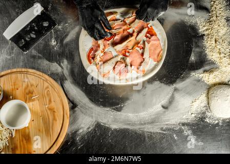 Pizza auf einem Stahltisch kochen; Teig, Mehl, Pizza-Belag, Sauce und Hände des Küchenchefs in schwarzen Handschuhen auf dem Tisch Stockfoto